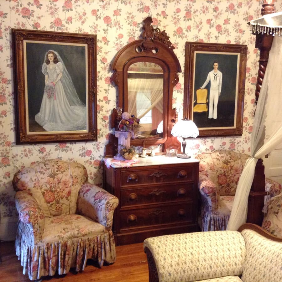 A vintage room featuring floral wallpaper, two armchairs, a wooden dresser, and framed portraits of a bride and a naval officer.