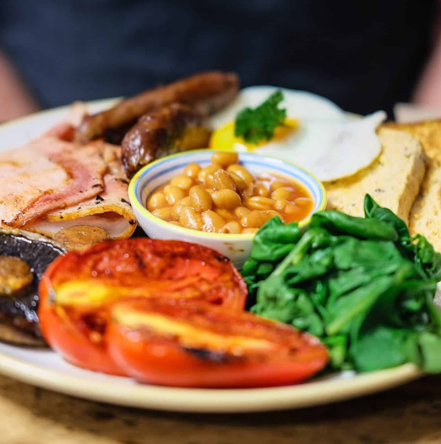 A hearty breakfast plate featuring sausage, bacon, baked beans, egg, grilled tomatoes, sautéed mushrooms, toast, and spinach.