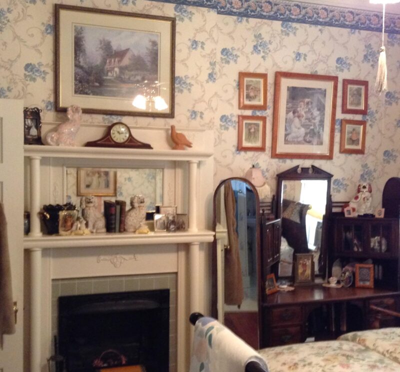 A cozy room featuring floral wallpaper, a vintage fireplace mantel, various framed pictures, and a wooden dresser with a mirror.