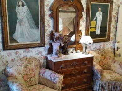 Antique furniture and floral wallpaper with two portraits hanging above a dresser.