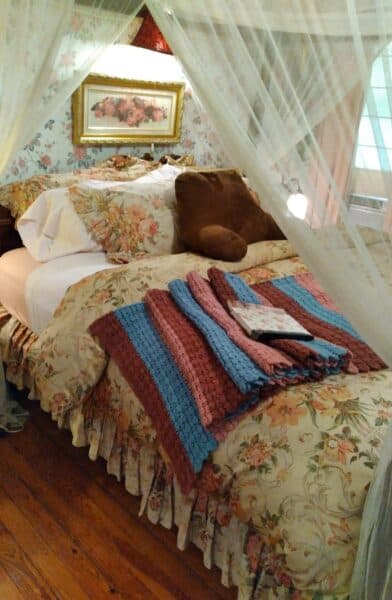 A cozy bed adorned with floral bedding, a plush pillow, and colorful knitted throws, all framed by delicate curtains.