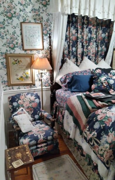 A cozy bedroom featuring floral wallpaper, a patterned bedspread, a lamp, and a matching chair.