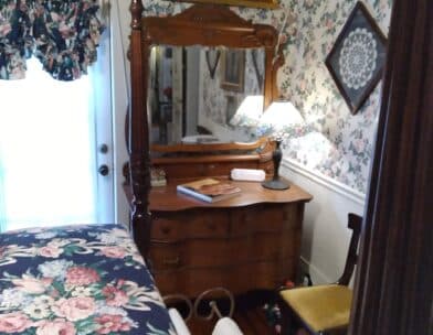 A cozy bedroom featuring a floral wallpaper, a wooden dresser with a mirror, and a soft lamp.