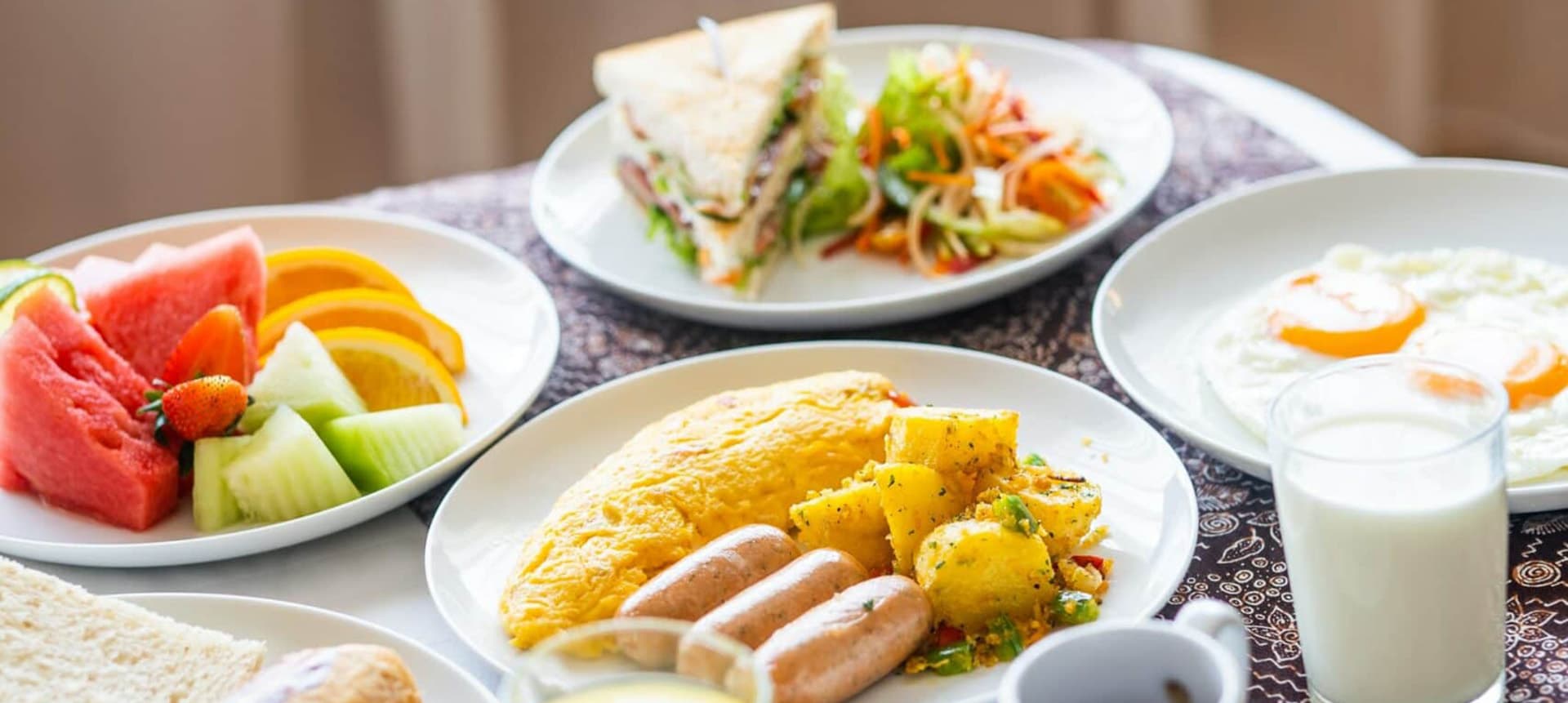 A colorful breakfast spread featuring fruit, omelette, sausages, a sandwich, salad, and eggs.